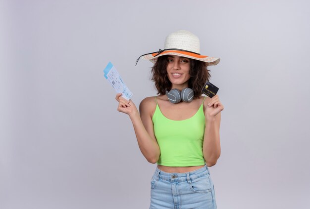 Una giovane donna felice con i capelli corti nella parte superiore del raccolto verde che porta il cappello del sole che mostra i biglietti aerei e la carta di credito su una priorità bassa bianca