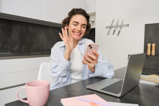 Una giovane donna felice dice ciao, agita la mano allo smartphone e sorride, chatta video con qualcuno sul cellulare.