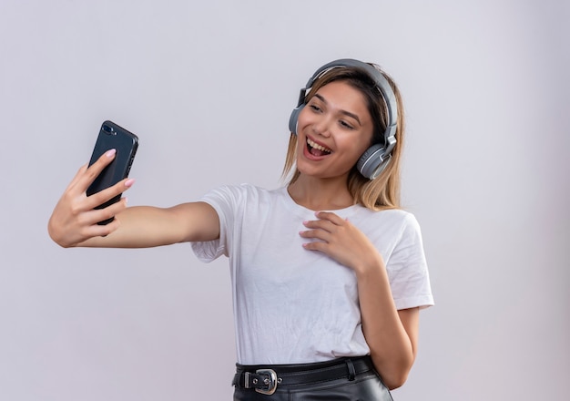 Una giovane donna felice in maglietta bianca che indossa le cuffie prendendo un selfie con lo smartphone su un muro bianco