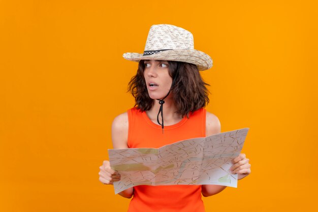 Una giovane donna frustrata con i capelli corti in una camicia arancione che porta la mappa della tenuta del cappello del sole e che osserva a destra per trovare il modo