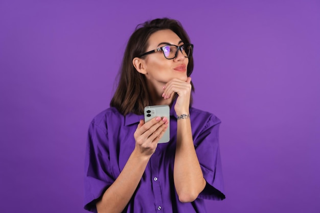 Una giovane donna in camicia e occhiali su uno sfondo viola con un telefono guarda pensierosa di lato con un leggero sorriso sulle labbra
