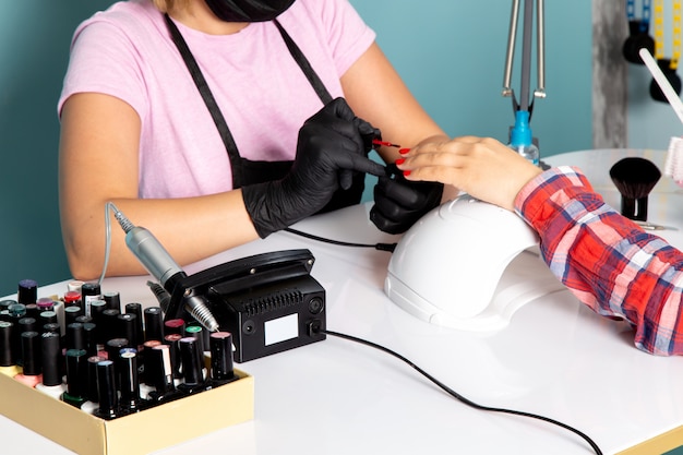 Una giovane donna manicure vista frontale in maglietta rosa con guanti neri e maschera nera che fa manicure sul blu