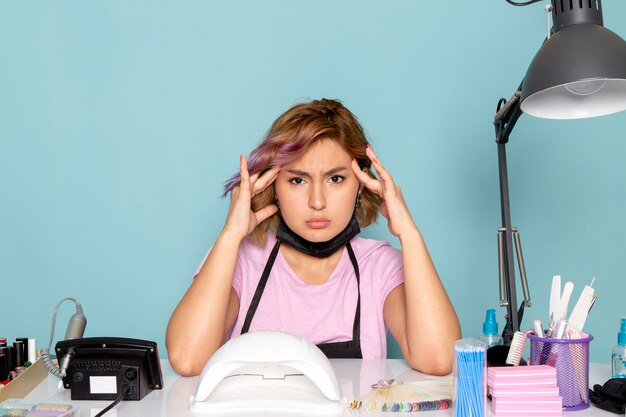 Una giovane donna manicure vista frontale in maglietta rosa con guanti neri e maschera nera con mal di testa sul blu