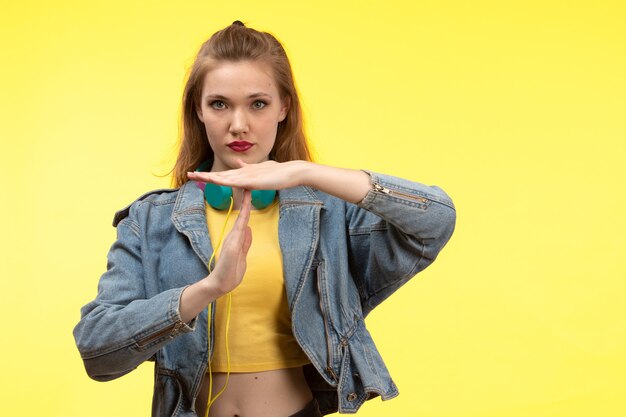 Una giovane donna moderna di vista frontale in pantaloni neri camicia nera e cappotto jean con auricolari colorati ascoltando musica in posa mostrando segno