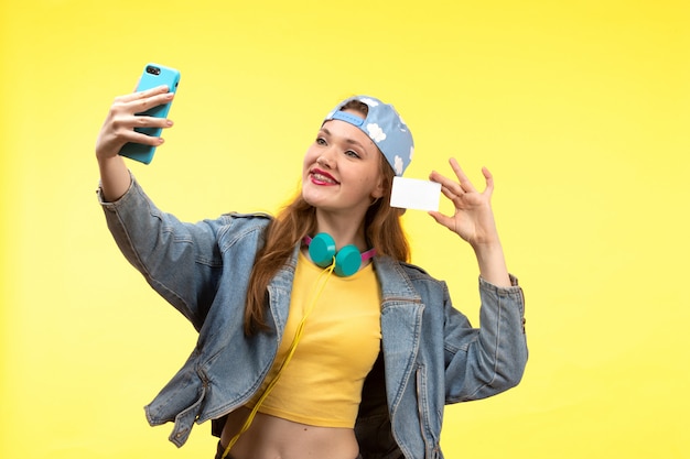 Una giovane donna moderna di vista frontale in pantaloni neri della camicia gialla e cappotto jean con le cuffie colorate che tengono carta bianca facendo uso del telefono che prende posa del selfie