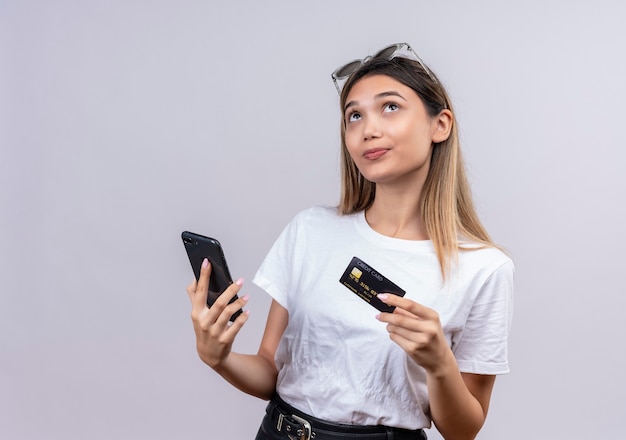 Una giovane donna premurosa in maglietta bianca in occhiali da sole pensando mentre si tiene il telefono cellulare e la carta di credito