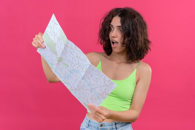 Una giovane donna scioccata con i capelli corti in top corto verde che guarda sorprendentemente la mappa con la bocca aperta