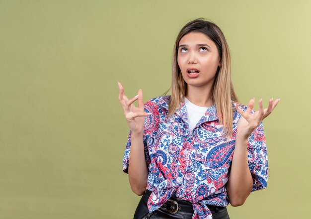 Una giovane donna scontenta che indossa una camicia stampata paisley alzando le mani mentre guarda su un muro verde
