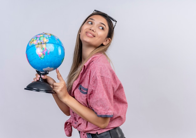 Una giovane donna soddisfatta che indossa una camicia rossa che punta a un globo mentre guarda il lato su un muro bianco