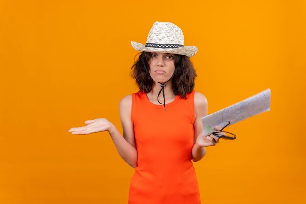 Una giovane donna sorprendente con i capelli corti in una maglietta arancione che indossa un cappello da sole che tiene la mappa non sapendo dove andare