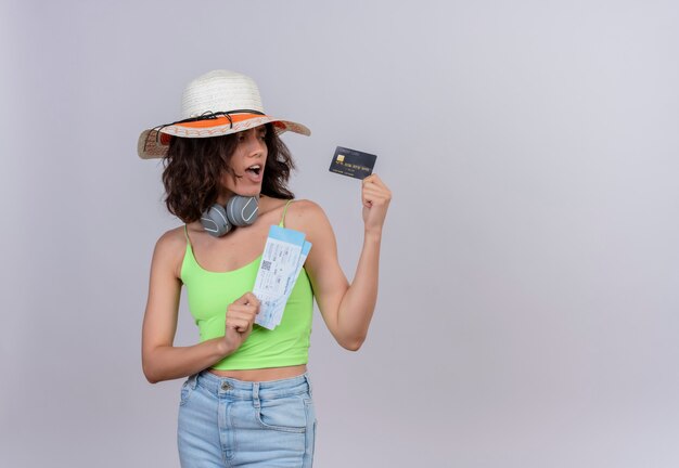 Una giovane donna sorpresa con i capelli corti nella parte superiore del raccolto verde in cuffie che portano il cappello del sole che tiene i biglietti aerei che esaminano la carta di credito su una priorità bassa bianca