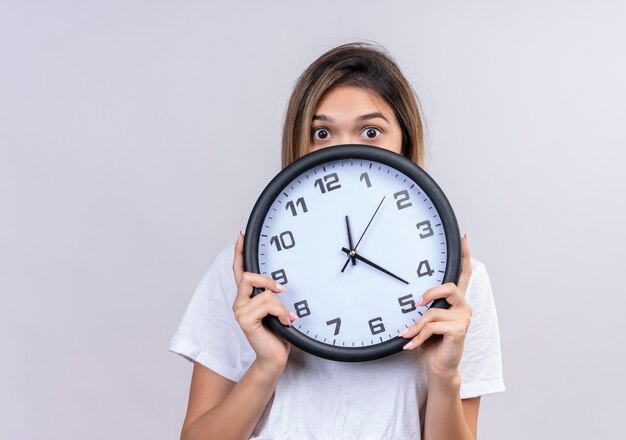 Una giovane donna sorpresa in t-shirt bianca che tiene orologio da parete mentre guarda