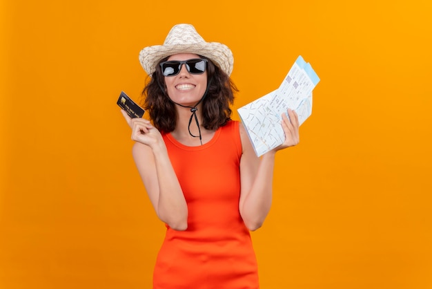 Una giovane donna sorridente con i capelli corti in una camicia arancione che indossa cappello da sole e occhiali da sole in possesso di mappa e carta di credito