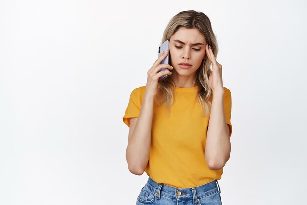 Una giovane donna turbata riceve una brutta telefonata, tiene lo smartphone vicino all'orecchio, accigliata turbata, in piedi sul bianco.