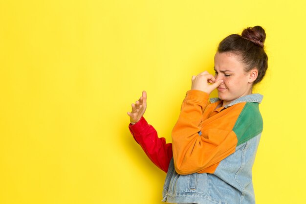 Una giovane donna vista frontale in giacca colorata camicia gialla e blue jeans tenendo il naso a causa dell'odore puzzolente