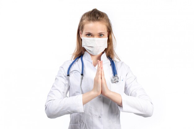 Una giovane dottoressa in vista frontale bianco in tuta medica bianca con stetoscopio indossando maschera protettiva bianca in posa mostrando pregare segno sul bianco