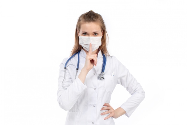 Una giovane dottoressa in vista frontale bianco in tuta medica bianca con stetoscopio indossando maschera protettiva bianca in posa segno di silenzio sul bianco