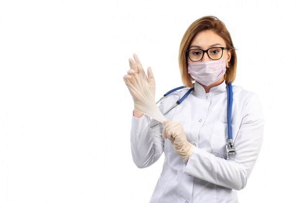 Una giovane dottoressa in vista frontale bianco vestito medico con stetoscopio indossando guanti protettivi maschera bianca sul bianco