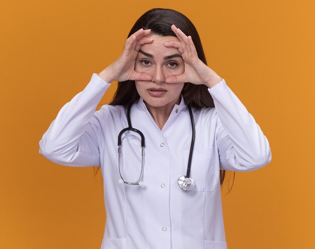 Una giovane dottoressa infastidita che indossa un abito medico con uno stetoscopio mette le mani sul viso isolato sulla parete arancione con spazio per le copie