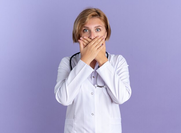 Una giovane dottoressa spaventata che indossa una tunica medica con uno stetoscopio ha coperto la bocca con la mano isolata su sfondo blu