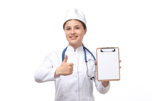 Una giovane dottoressa vista frontale in abito medico bianco con stetoscopio blu tenendo il blocco note l