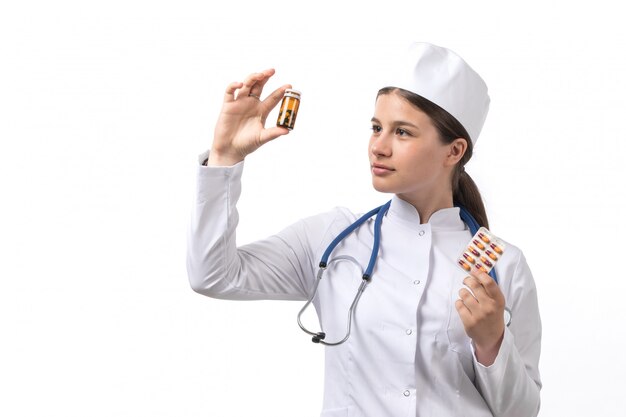 Una giovane dottoressa vista frontale in tuta medica bianca e berretto bianco con stetoscopio blu tenendo pillole e pozioni