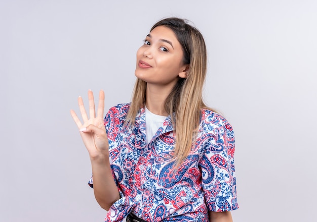 Una giovane e bella donna che indossa una camicia stampata paisley che mostra il numero quattro con le dita mentre guarda su un muro bianco