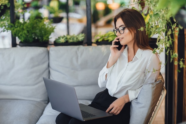 Una giovane e bella donna lavora su un laptop sulla terrazza estiva del suo ufficio moderno e parla al telefono