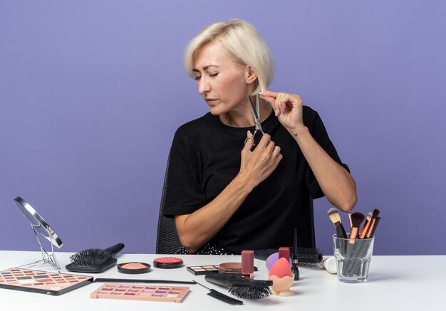 Una giovane e bella ragazza scontenta si siede al tavolo con strumenti per il trucco che tagliano i capelli con le forbici isolate su sfondo blu