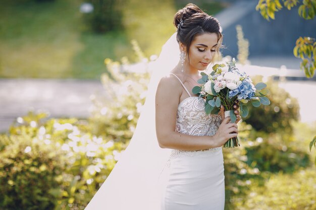 Una giovane e bella sposa è in piedi in un parco estivo con bouquet di fiori