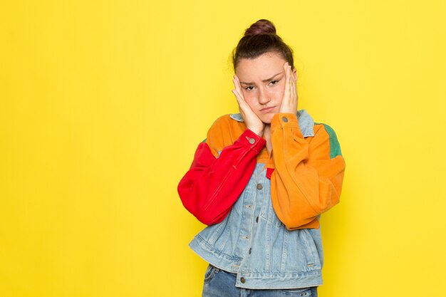 Una giovane femmina di vista frontale in giacca colorata e blue jeans della camicia gialla che posano con l'espressione depressa