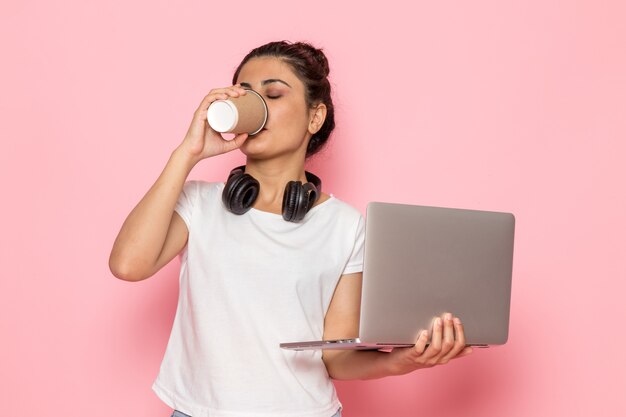 Una giovane femmina di vista frontale in maglietta e blue jeans bianche facendo uso del computer portatile e bevendo caffè