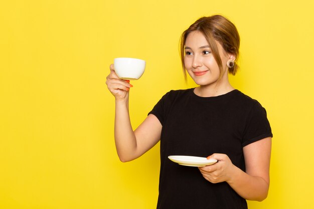 Una giovane femmina di vista frontale in vestito nero che beve caffè e che sorride sul giallo