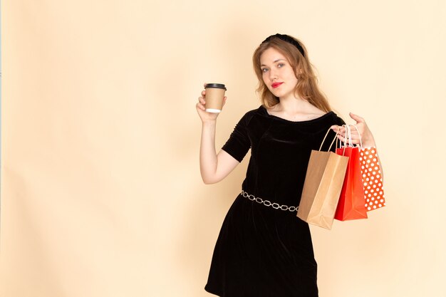 Una giovane femmina di vista frontale in vestito nero con la cinghia della catena che tiene i pacchetti della spesa e la tazza di caffè sul beige