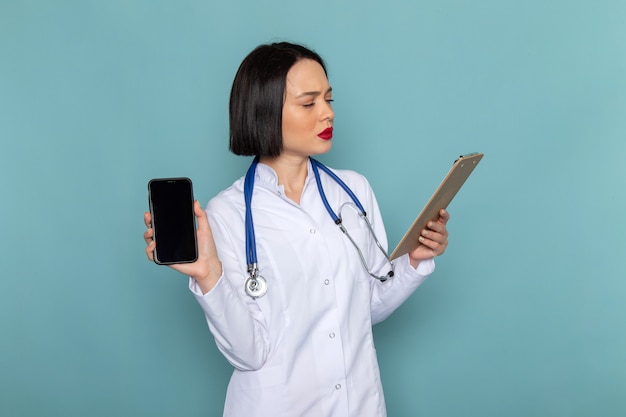 Una giovane infermiera femminile di vista frontale in tuta medica bianca e stetoscopio blu con blocco note e telefono