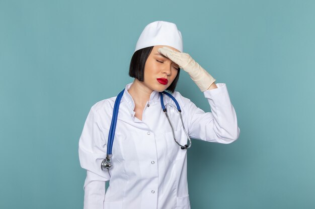 Una giovane infermiera femminile di vista frontale in tuta medica bianca e stetoscopio blu con mal di testa