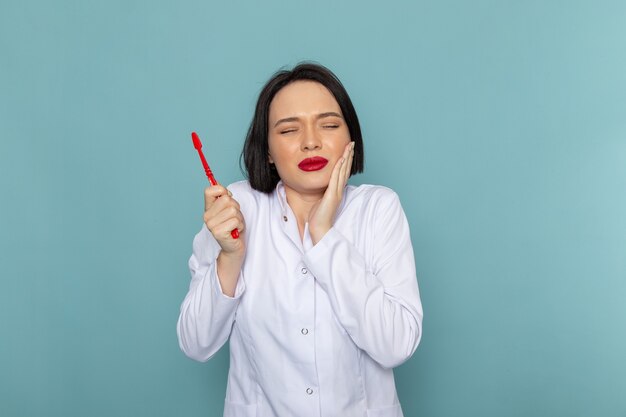 Una giovane infermiera femminile di vista frontale in vestito medico bianco che ha un mal di denti sul medico dell'ospedale della medicina dello scrittorio blu