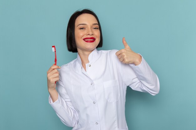 Una giovane infermiera femminile di vista frontale in vestito medico bianco che sorride tenendo lo spazzolino da denti sul medico dell'ospedale della medicina dello scrittorio blu