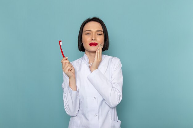 Una giovane infermiera femminile di vista frontale in vestito medico bianco che tiene lo spazzolino da denti sul medico dell'ospedale della medicina dello scrittorio blu