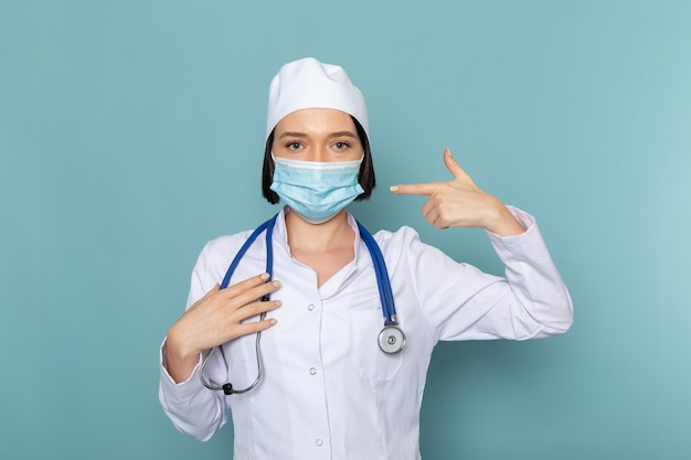 Una giovane infermiera femminile di vista frontale in vestito medico bianco e maschera da portare dello stetoscopio blu sul medico dell'ospedale della medicina dello scrittorio blu