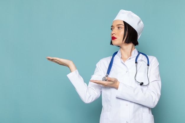 Una giovane infermiera femminile di vista frontale in vestito medico bianco e stetoscopio blu che posa sul medico blu dell'ospedale della medicina dello scrittorio