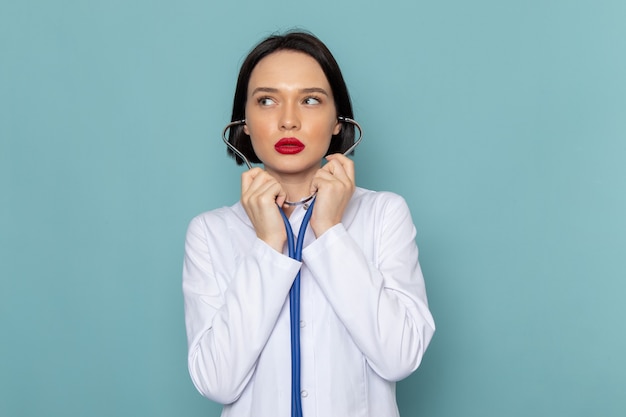 Una giovane infermiera femminile di vista frontale in vestito medico bianco e stetoscopio blu che posa sul medico blu dell'ospedale della medicina dello scrittorio