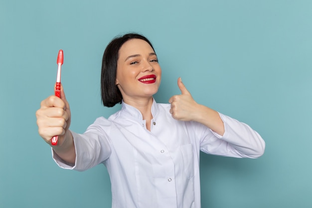 Una giovane infermiera femminile di vista frontale in vestito medico bianco e stetoscopio blu che tiene lo spazzolino da denti sul medico dell'ospedale della medicina dello scrittorio blu