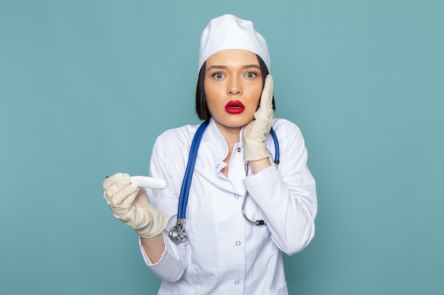 Una giovane infermiera femminile di vista frontale in vestito medico bianco e stetoscopio blu con l'espressione sorpresa sul vestito blu di medico dell'ospedale della medicina dello scrittorio