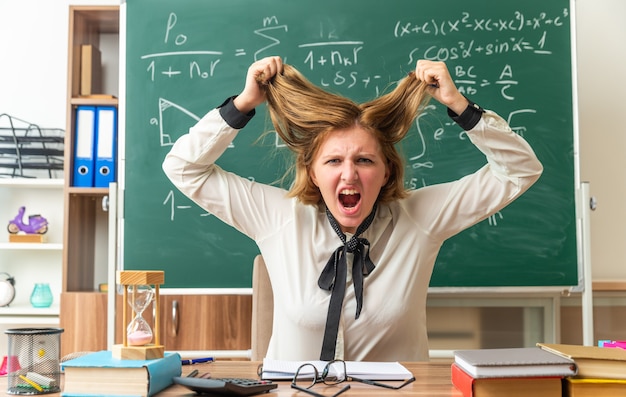 una giovane insegnante arrabbiata si siede al tavolo con materiale scolastico afferrato i capelli in classe