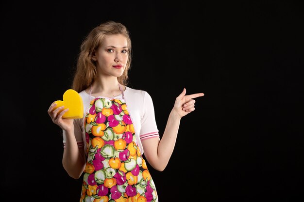 Una giovane ragazza attraente di vista frontale in capo variopinto che tiene forma gialla del cuore sulla positività nera di sorriso di amore del fondo