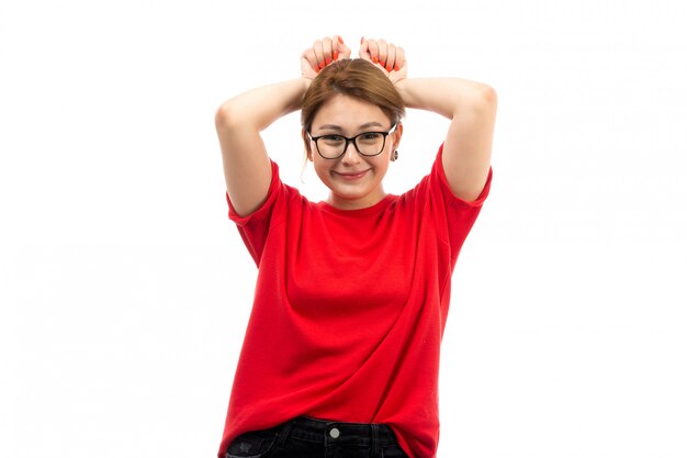 Una giovane ragazza attraente di vista frontale in maglietta rossa che porta i jeans neri in occhiali da sole che posano civetta sorridente sul bianco