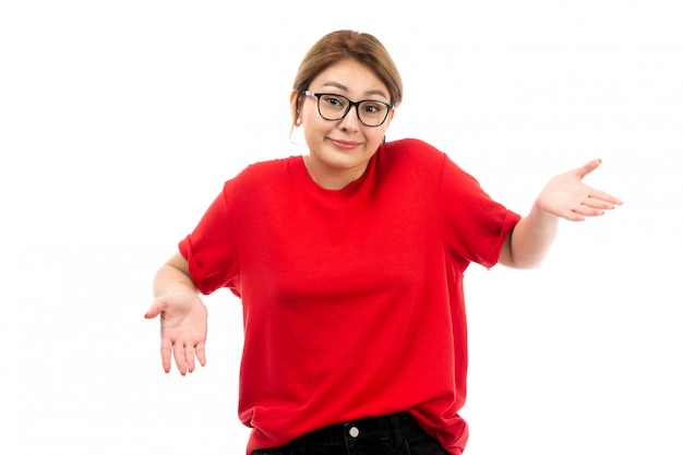 Una giovane ragazza attraente di vista frontale in maglietta rossa che porta i jeans neri in occhiali da sole che posano sul bianco
