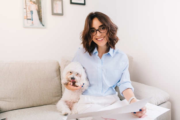 Una giovane ragazza bruna con una camicia blu è seduta sul divano in studio. Abbraccia il cagnolino bianco e tiene alcuni schizzi sulle ginocchia. Sta sorridendo alla telecamera.