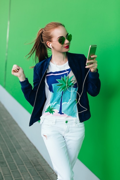 Una giovane ragazza con una giacca blu e pantaloni bianchi è in piedi all'aperto vicino al muro verde con una linea bianca verso il basso. La ragazza indossa occhiali da sole con cuori. Sta alleggerendo la musica con l'aiuto delle cuffie.